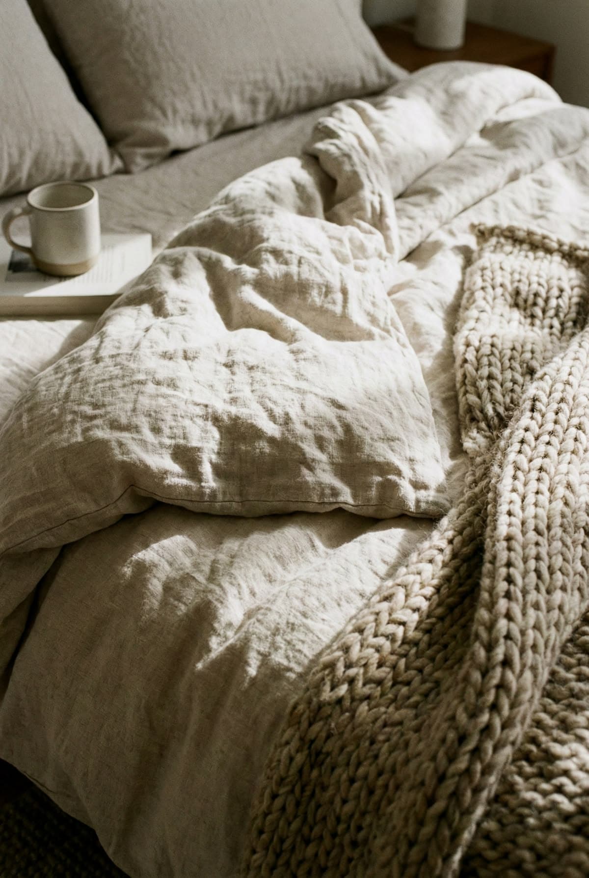 A bed layered in rumpled natural linen bedding with a chunky cream wool knit throw folded across it, a mug and an open book resting on the edge.