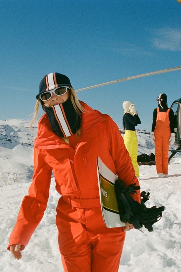 A skier in an orange snowsuit and a striped balaclava carrying a snowboard across a sunny mountain ridge, with two friends and a helicopter behind her.